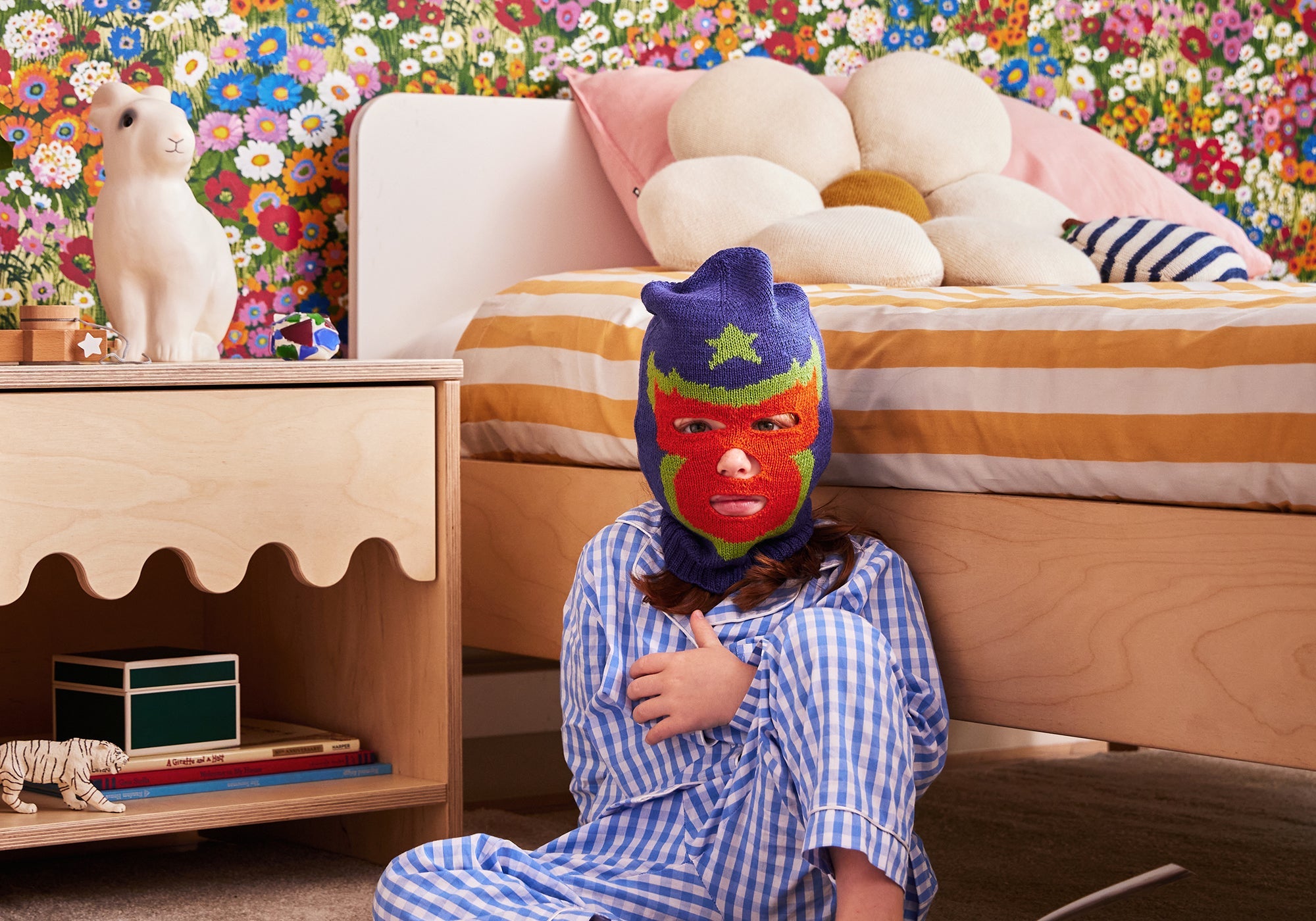 Child in blue pajamas wearing a colorful mask sits on the floor by a bed with floral wallpaper in the background.