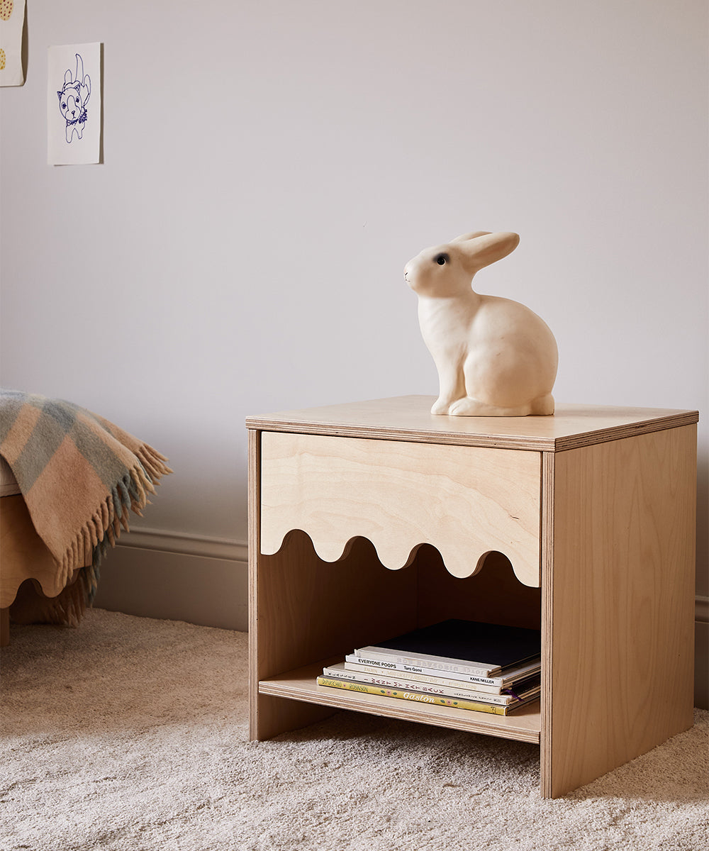 A wooden bedside table with wavy skirting showcases the Le Bunny Lamp atop, while a few books sit neatly on the lower shelf. A drawing decorates the wall above.