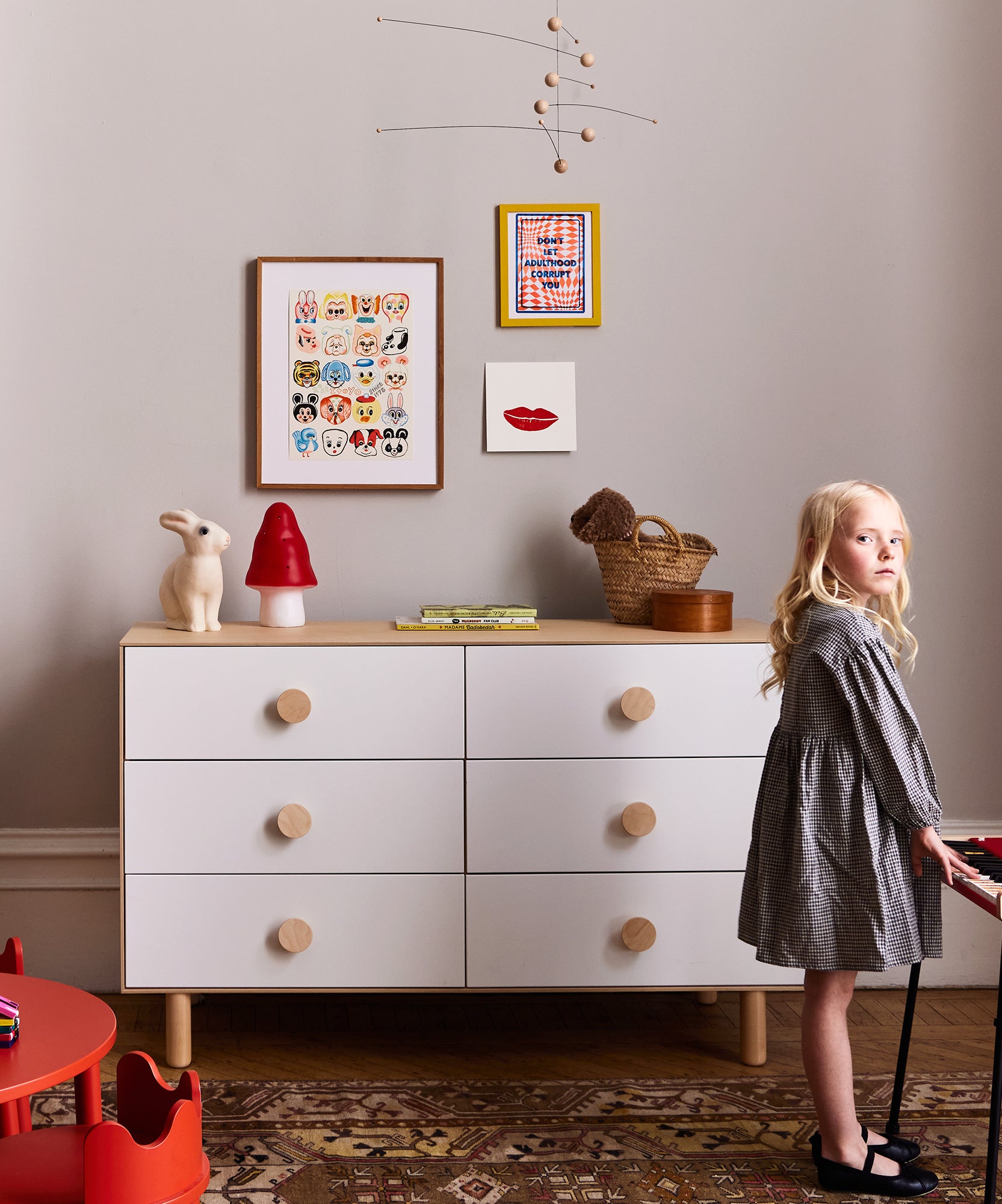 A young girl stands by a dresser with the Le Small Mushroom Lamp, art on the wall, and toys in a cozy, modern children's room.