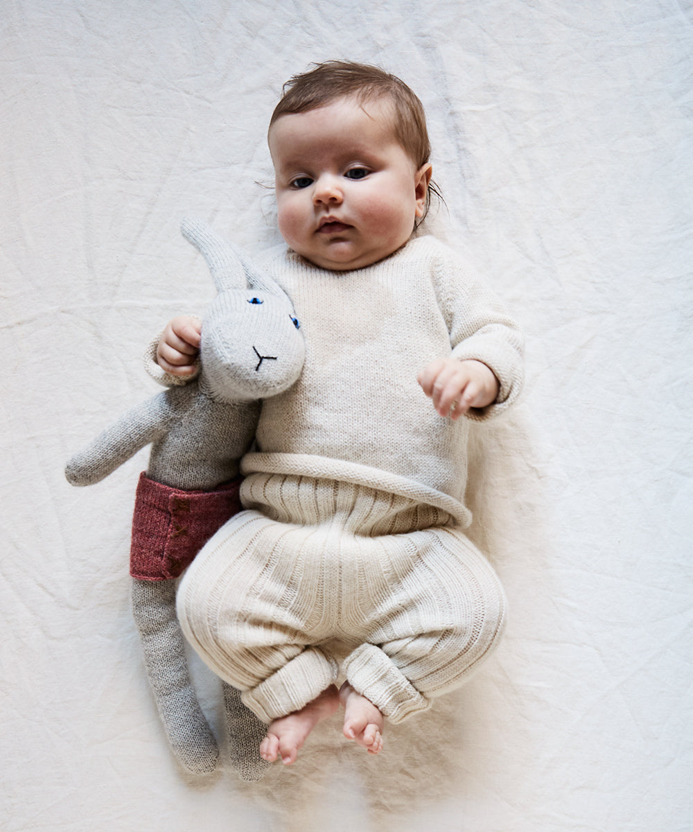 A baby in cream knitwear lies on a Crib/Toddler Fitted Sheet, holding a stuffed rabbit toy with red shorts.