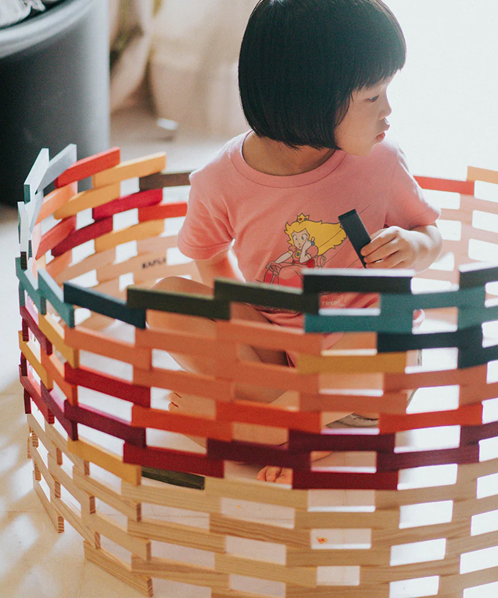 A young child in a pink shirt builds and sits inside a large circular structure using the KAPLA 100 Octocolor Case wooden blocks.