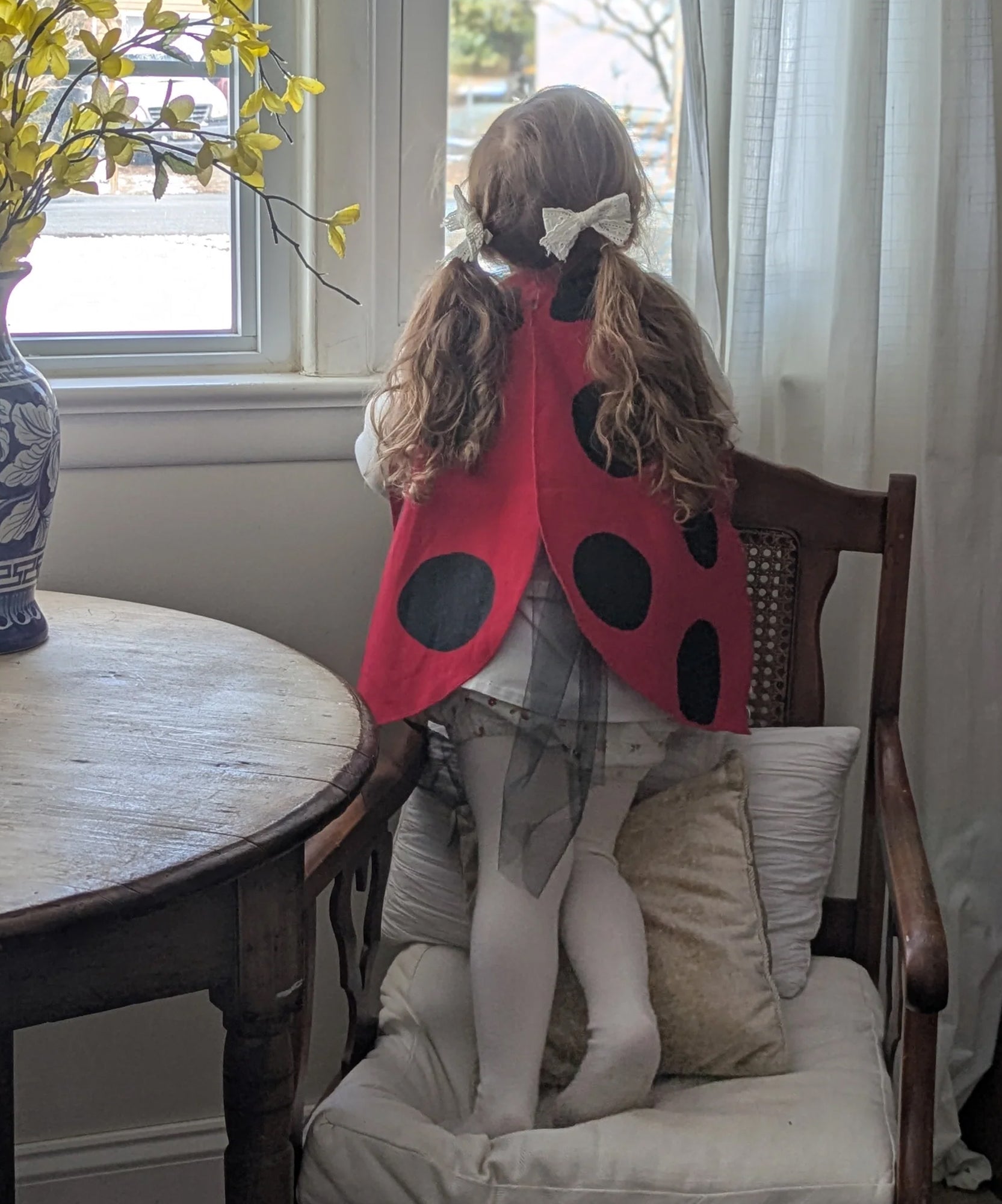 A child wearing Ladybug Wings stands on a chair, gazing out a window next to a round table with a vase.