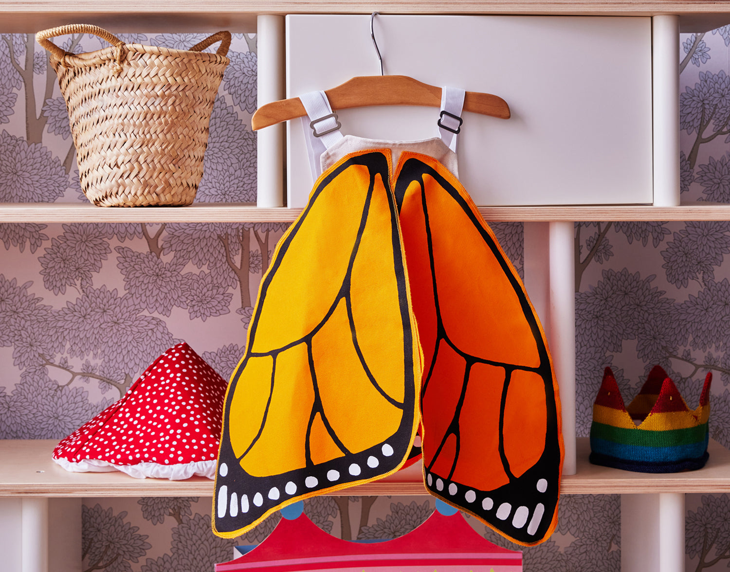 A butterfly wing costume hangs on a wooden hanger in a closet, with a straw basket, red polka dot hat, and rainbow crown on the shelves.