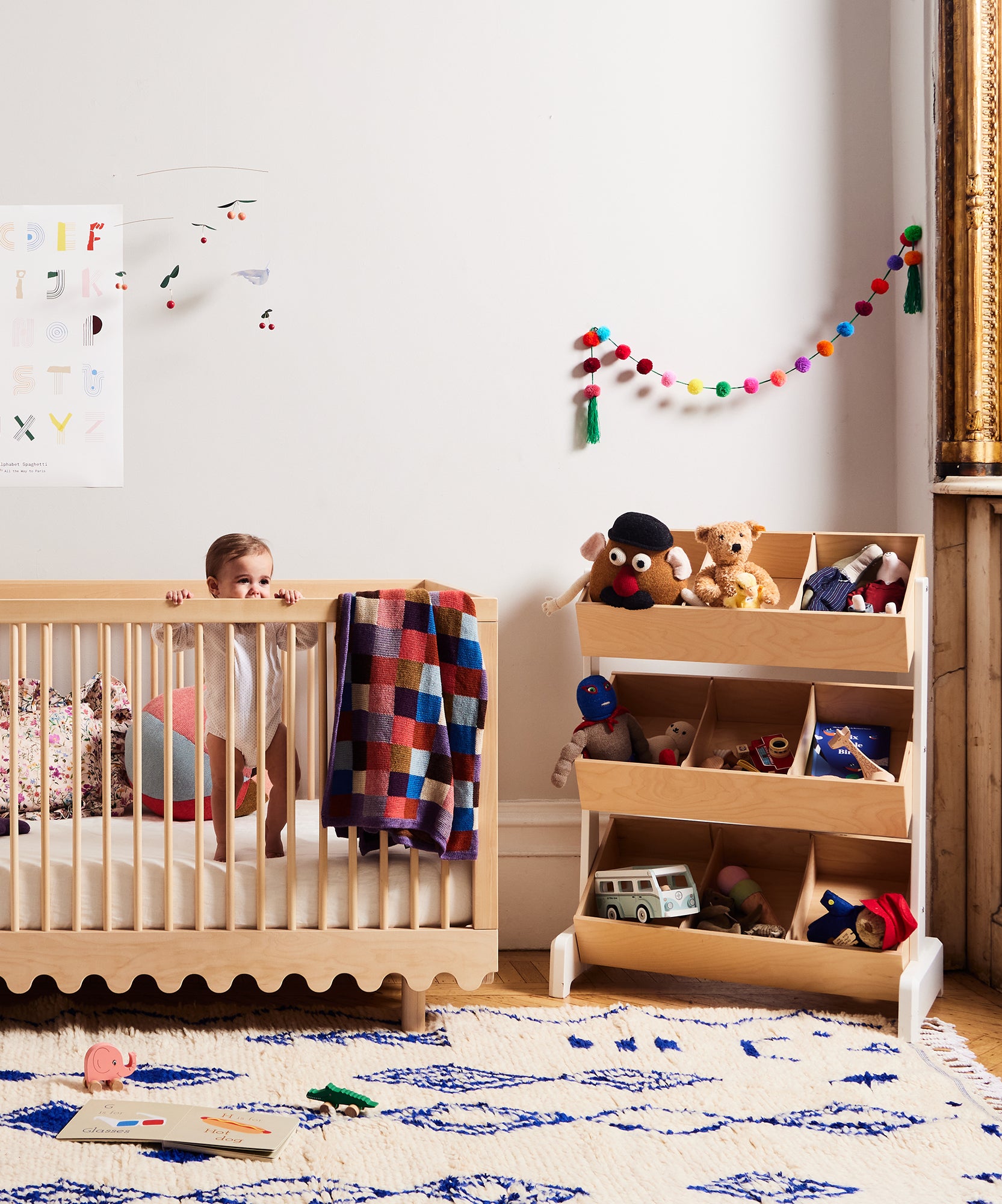A baby stands in a crib beside Toy Store toys, books, and a garland in a bright, playful nursery.