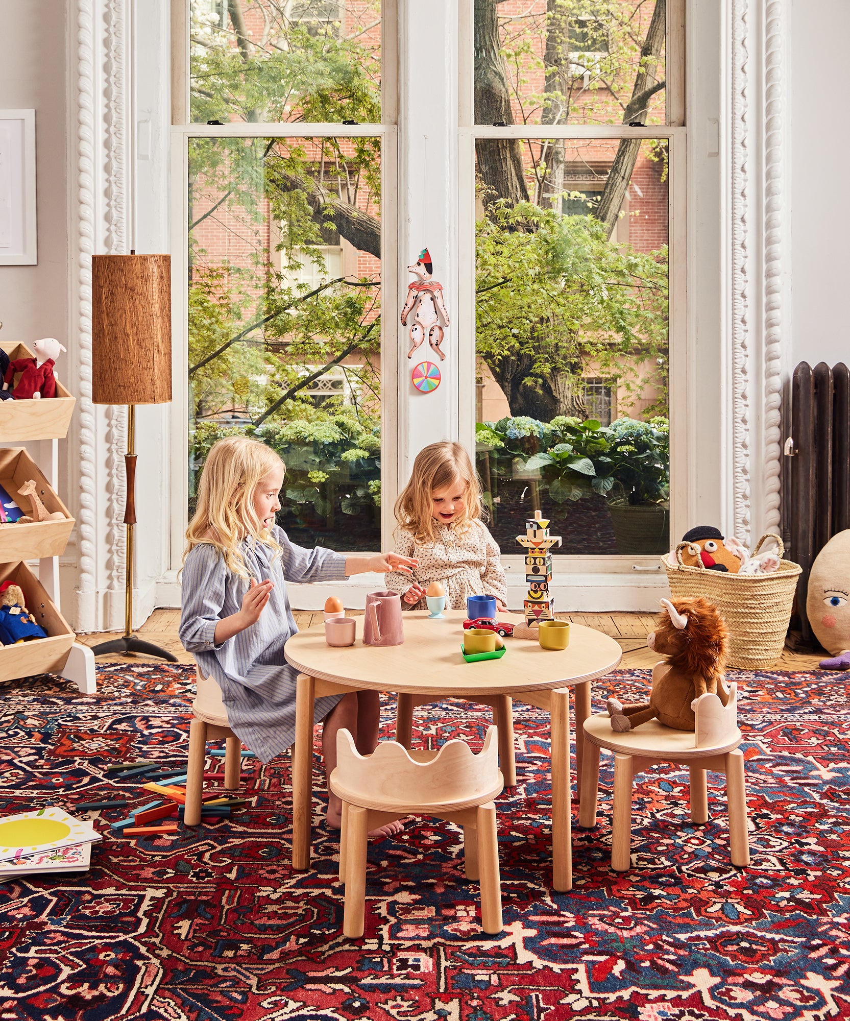 Two young children play with toys at a round Moss Table in a colorful, cozy room with a large window and patterned rug.