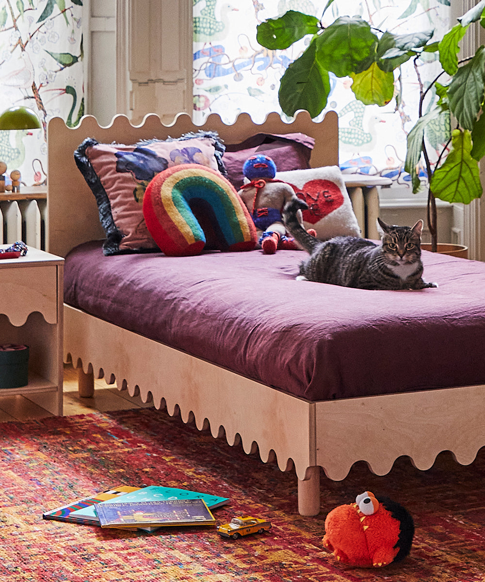 A cat lies on a purple bed with a Rainbow Pillow in a cozy, toy-filled bedroom.