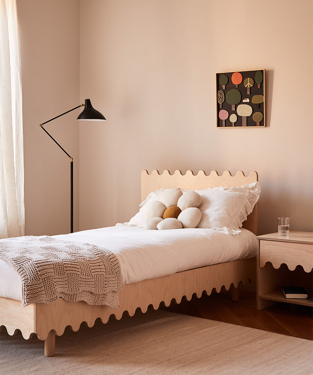 Minimalist bedroom with a wavy-edged bed, Daisy Pillow, and a standing lamp. Abstract art adorns the wall.
