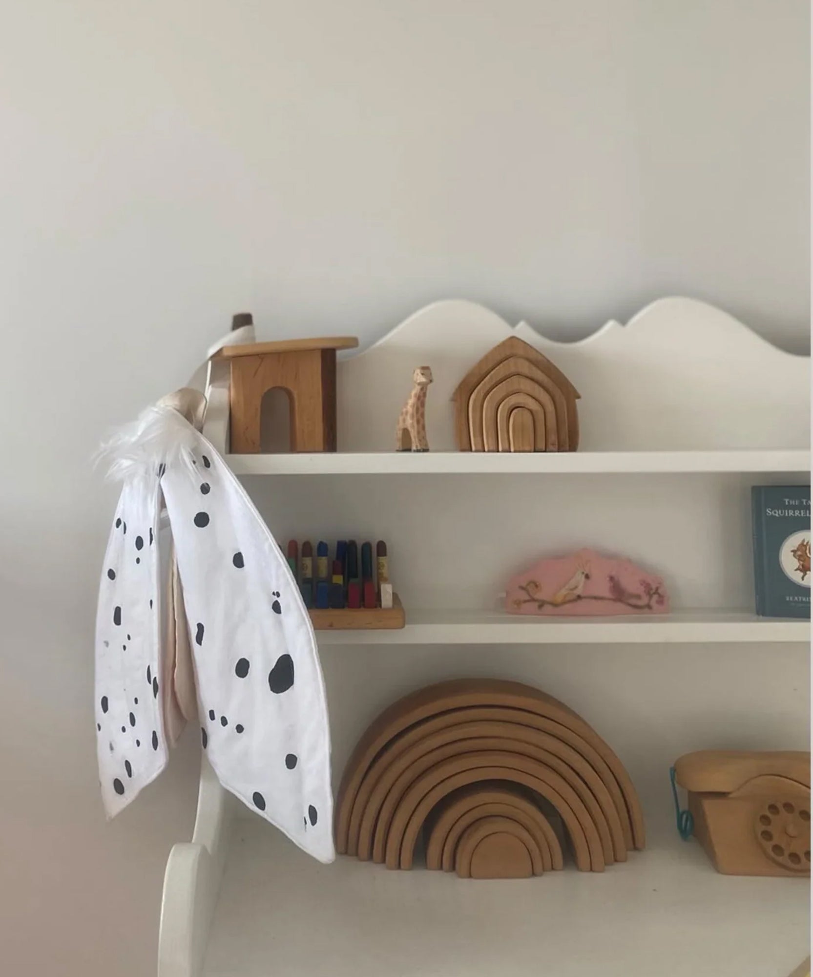 White shelf with wooden toys, stacked rainbows, a book, and Le Leopard Moth Wings cape against a plain wall.