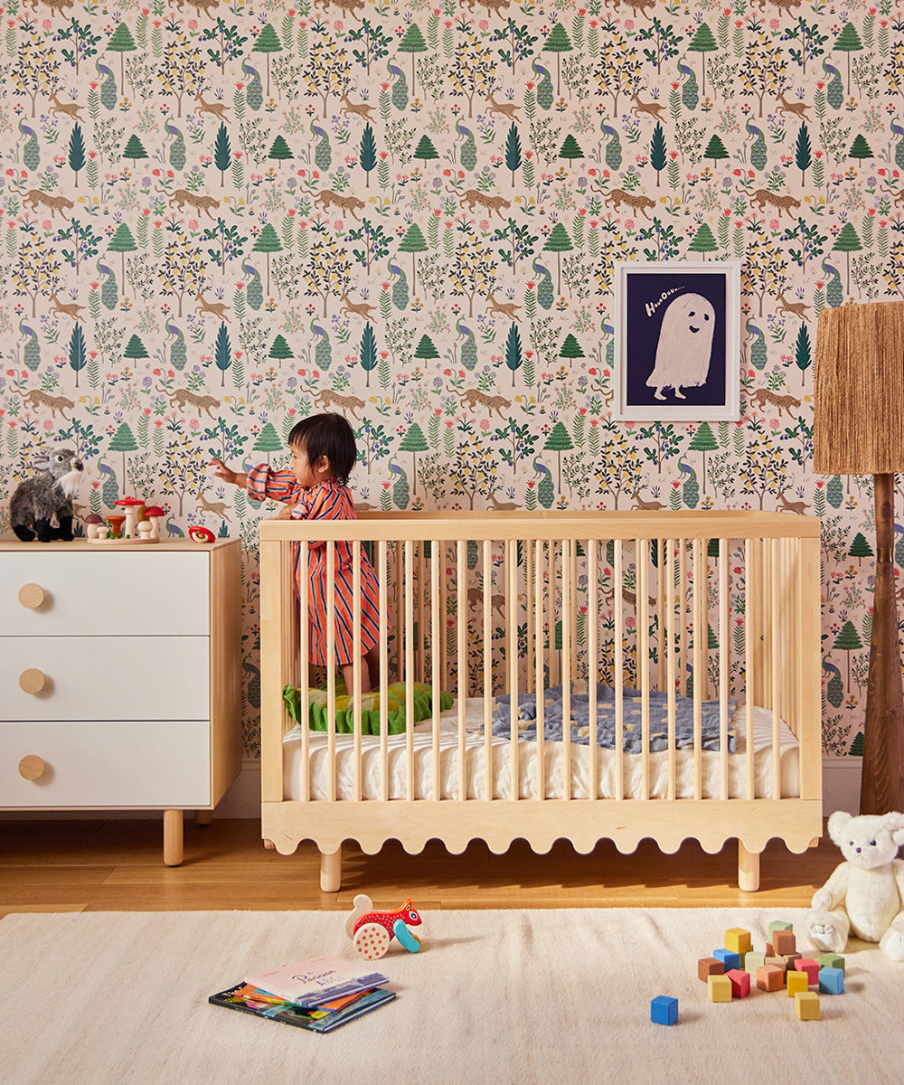 A toddler stands by a dresser next to a crib in a colorful nursery with patterned wallpaper and the Le Squirrel Toy on the floor.