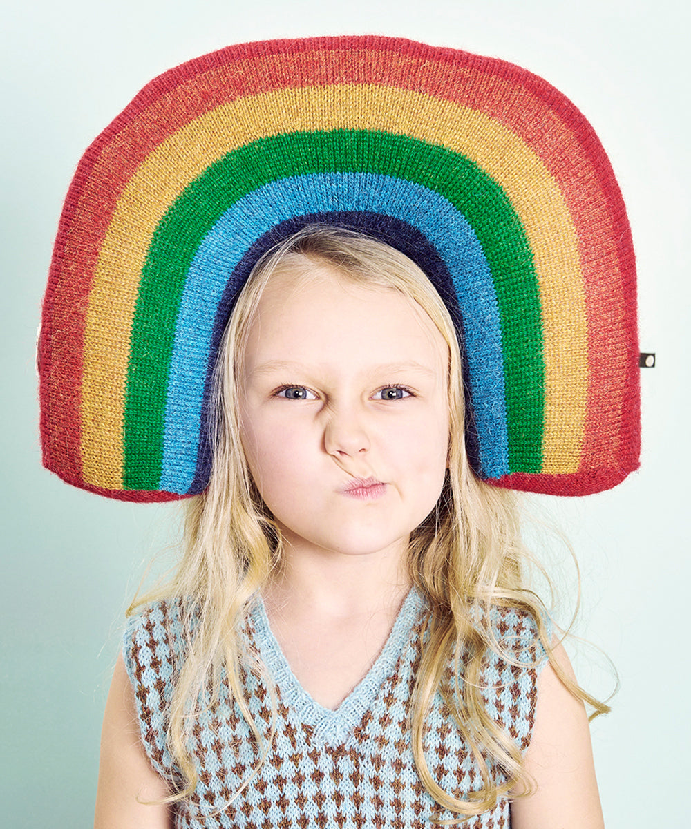 A young girl with long blonde hair makes a playful face while wearing the Rainbow Pillow as a vibrant headpiece.