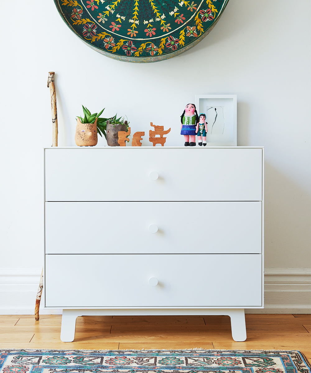 The Sparrow 3-Drawer Dresser, adorned with potted plants, figurines, and artwork on top, stands against a white wall.