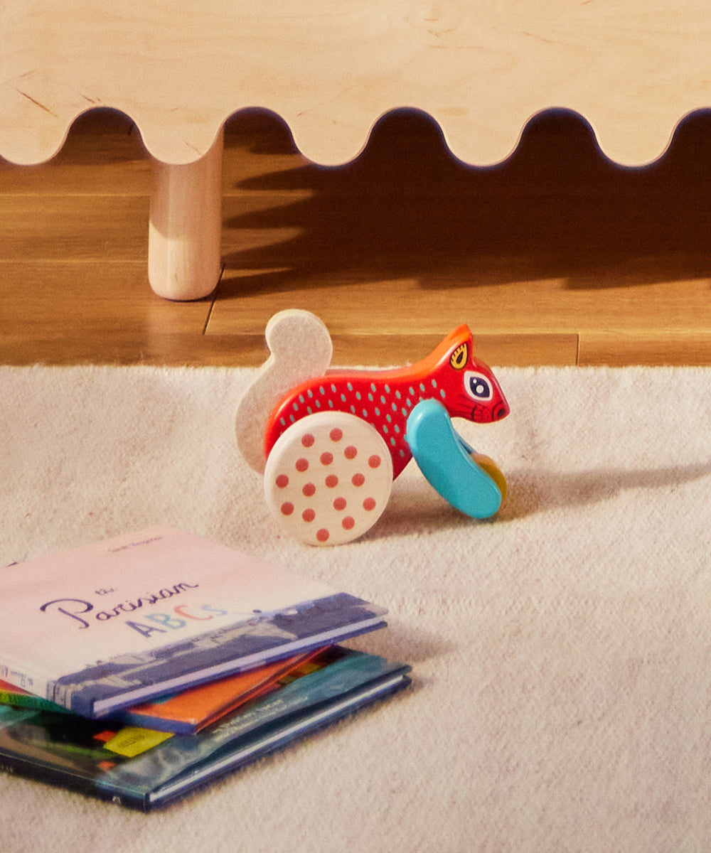 Le Squirrel Toy, a red wooden squirrel, sits on a rug near children's books under a wooden table.
