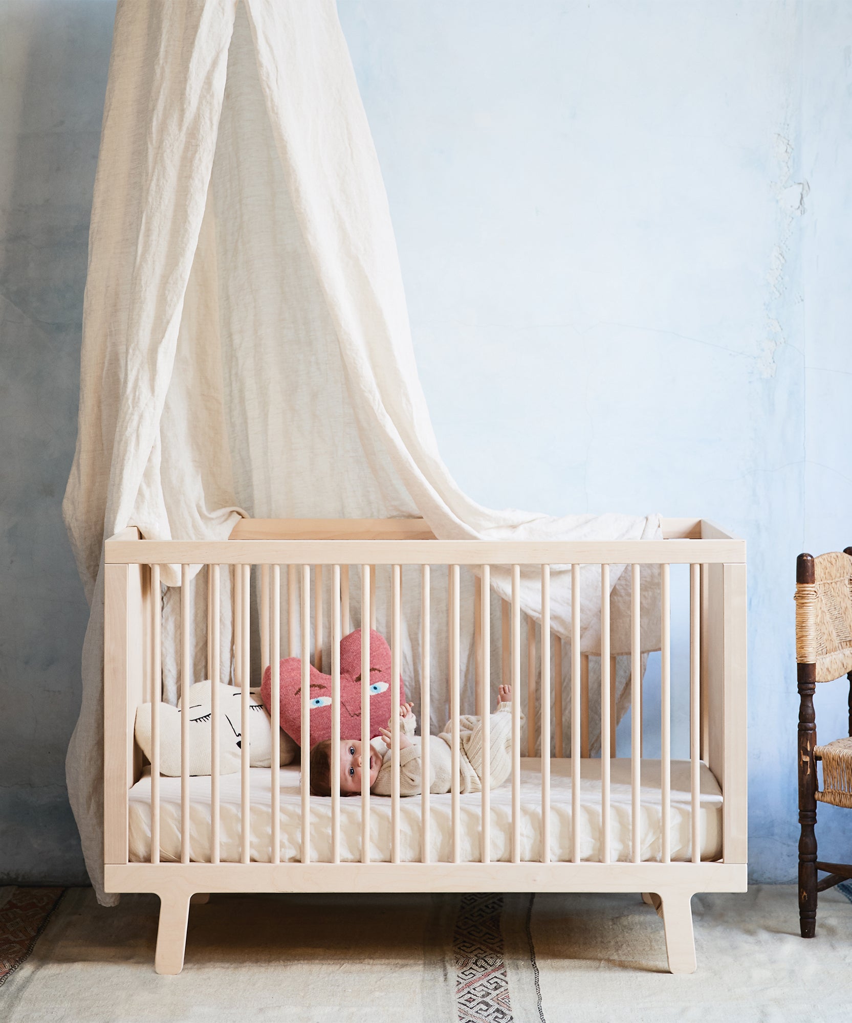 A baby rests in a wooden crib outfitted with the Crib/Toddler Fitted Sheet, under a white canopy, beside a rustic chair.