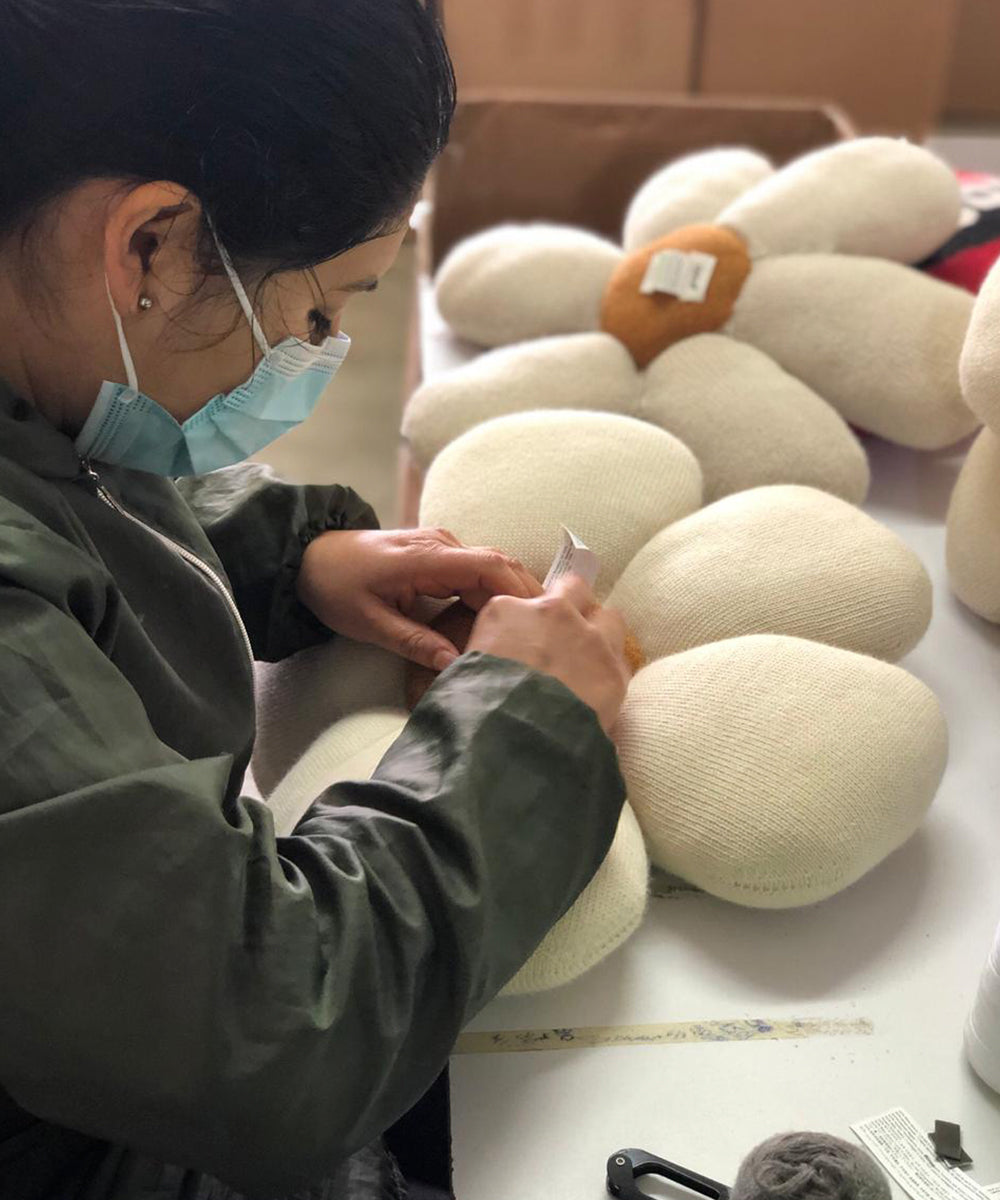 A person in a mask and green jacket sews a Daisy Pillow plush.