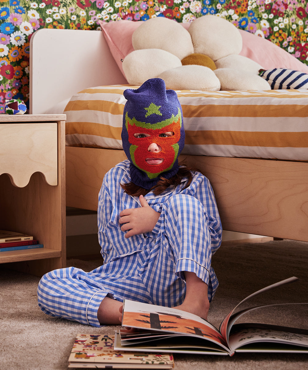 A child wearing a Luchador Mask and blue pajamas is sitting on the floor with books in a colorful room.