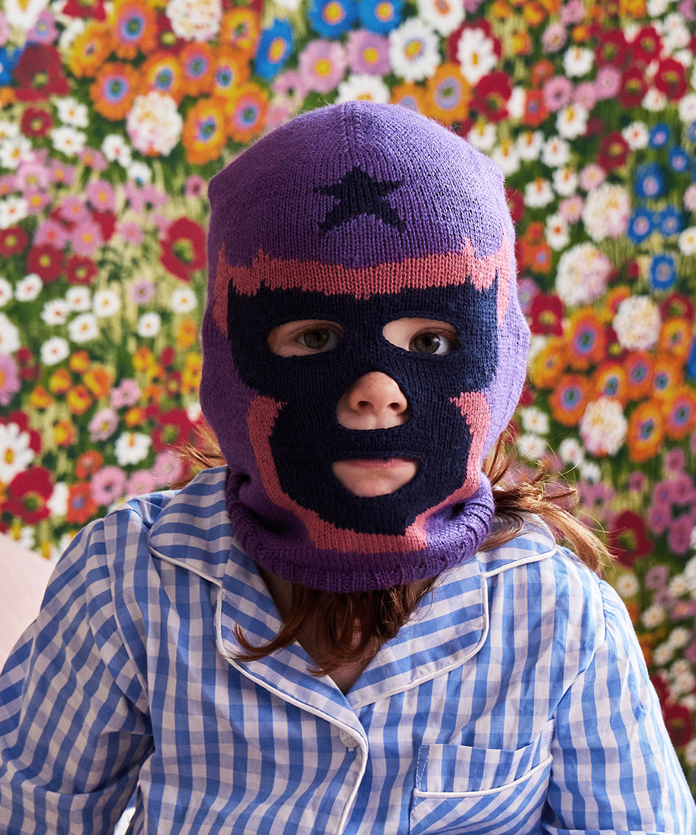 Child wearing a Luchador mask and checkered pajamas sits in front of a colorful floral backdrop.