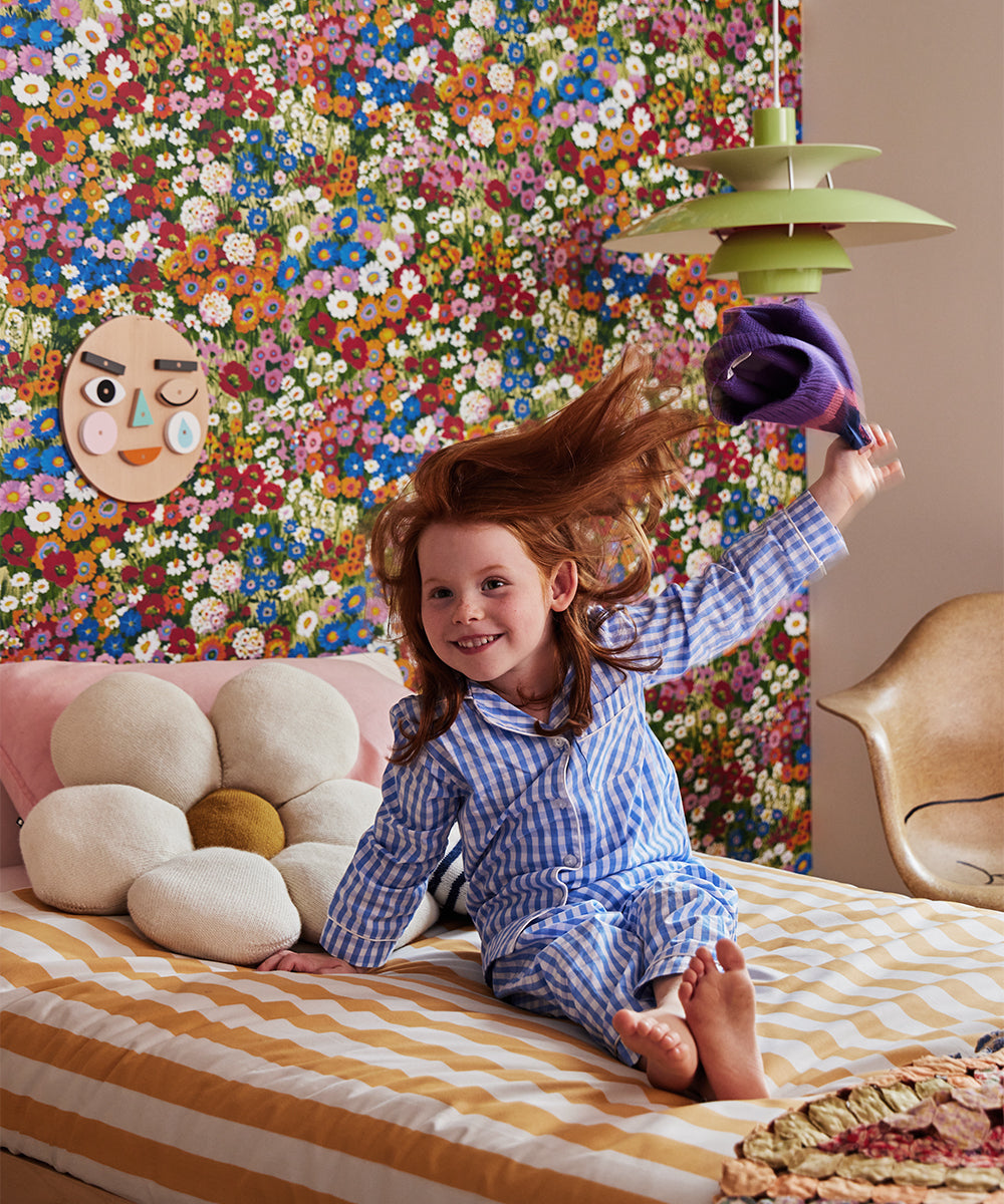Child wearing a luchador mask happily tossing a purple hat, sitting on a bed with vibrant flower wallpaper behind.