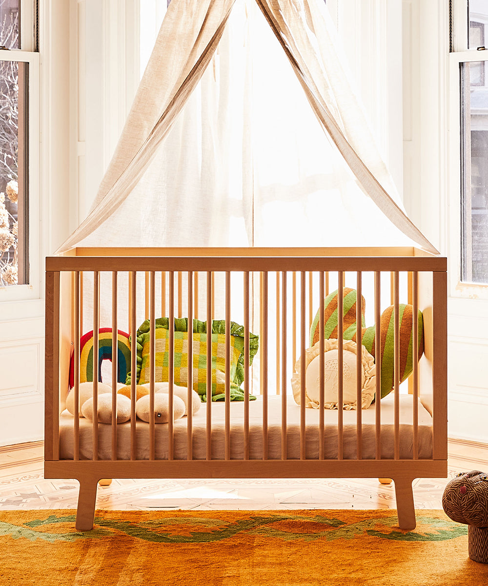 Light wood crib with a Rainbow Pillow and canopy, set in a bright, sunlit room with large windows.