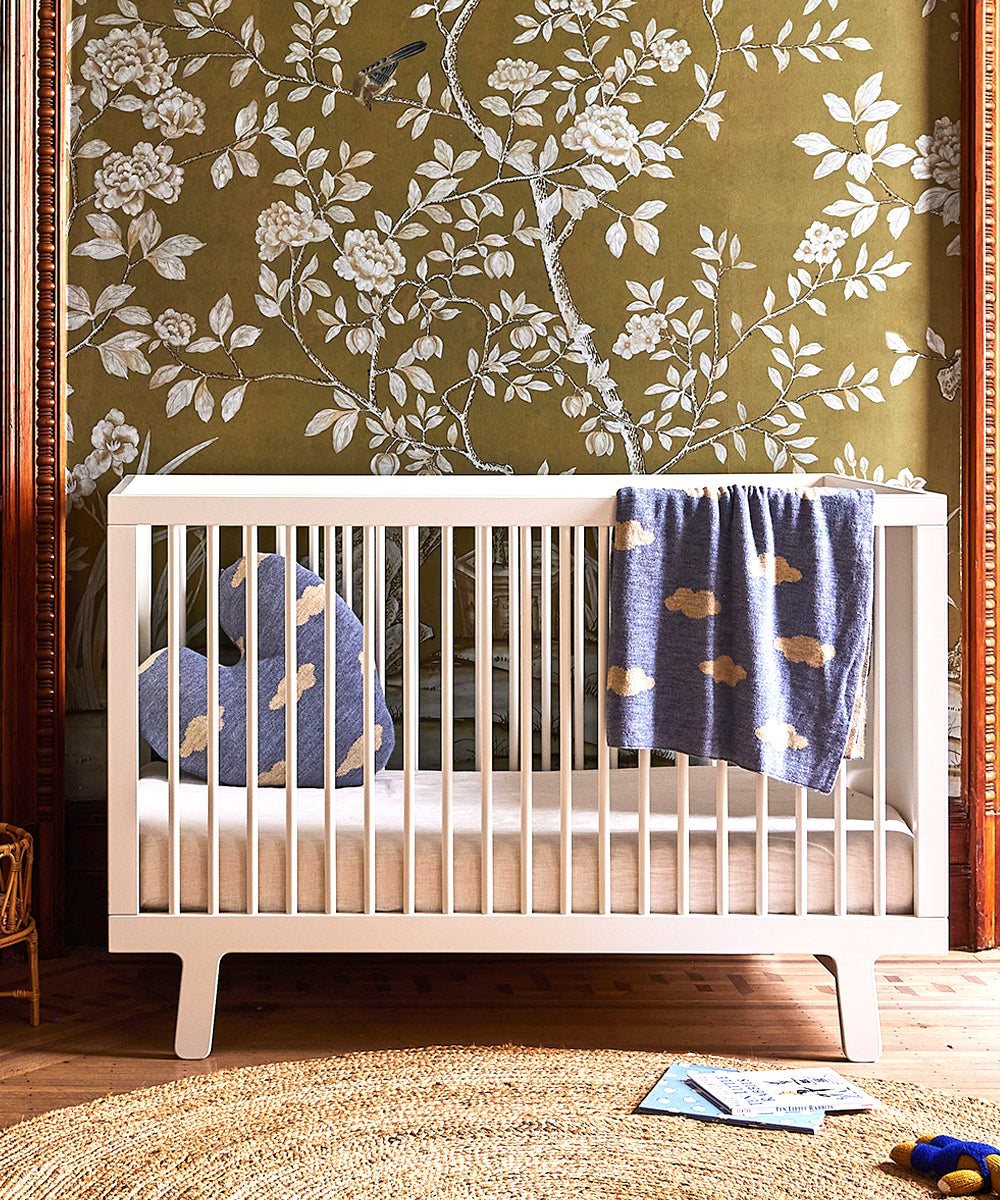 White crib with Heart Cloud Pillow and blanket, floral wallpaper, round rug, wooden floor, and toys on the side.