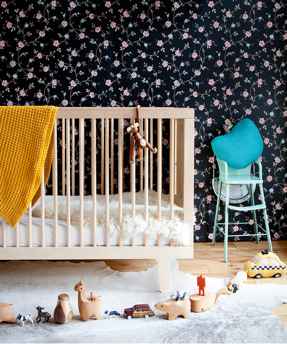 The nursery displays a GREENGUARD Gold certified Sparrow Crib made from birch hardwood, with a yellow blanket and toys on the floor. A high chair sits against floral wallpaper, creating a cozy retreat for your little one.