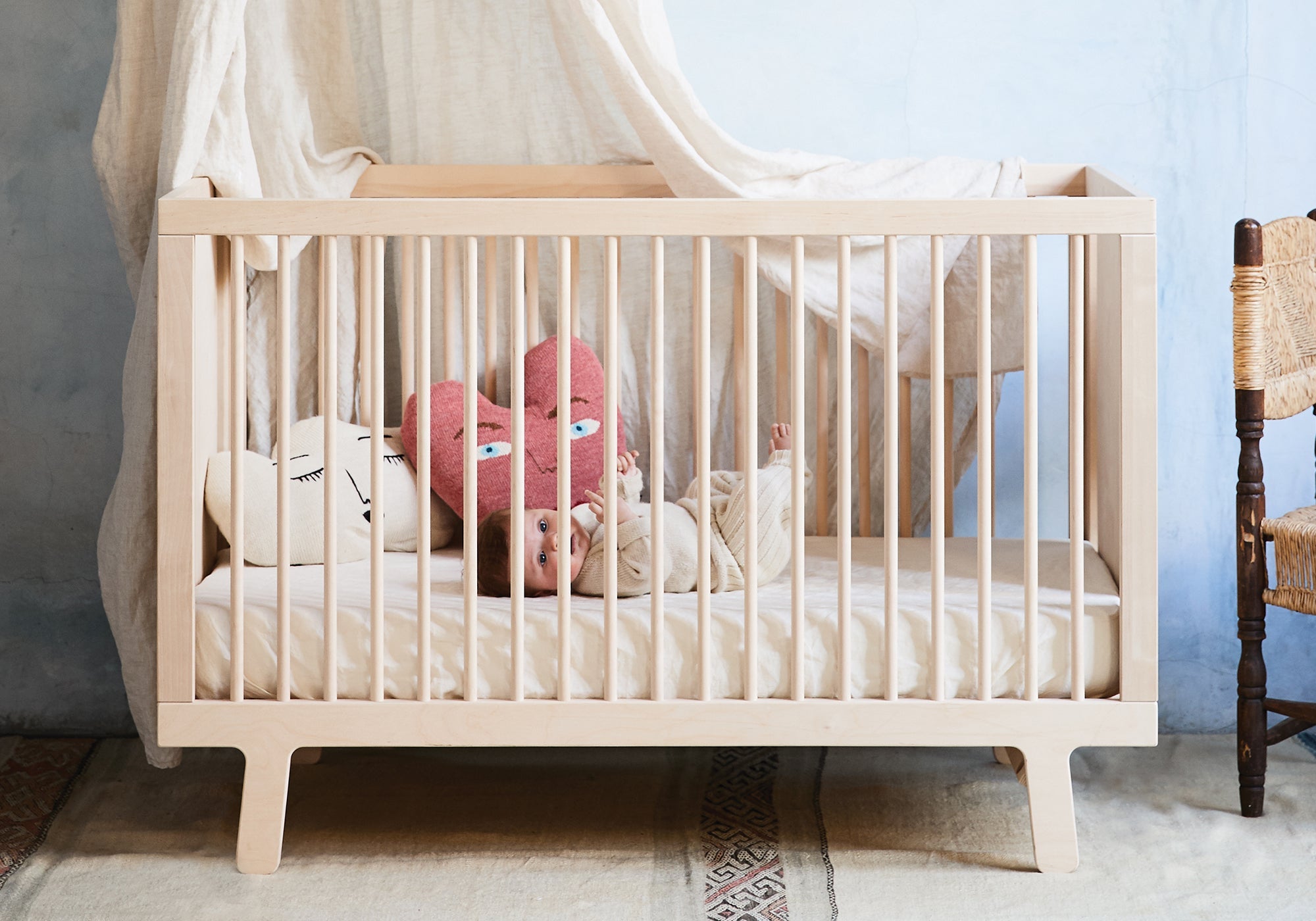 Baby lying in a light wooden crib with plush pillows and a canopy, next to a woven chair.