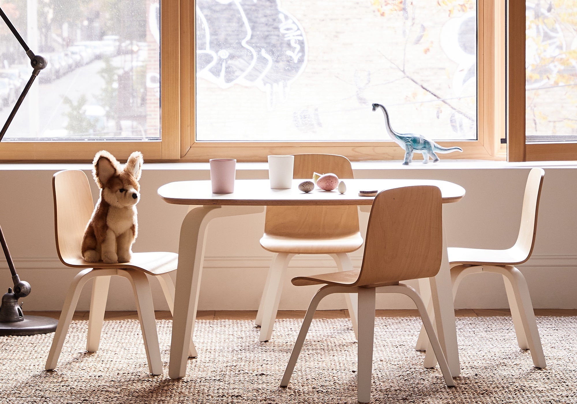 Child-sized table and chairs with plush toy and dinosaur figurine by a large window.