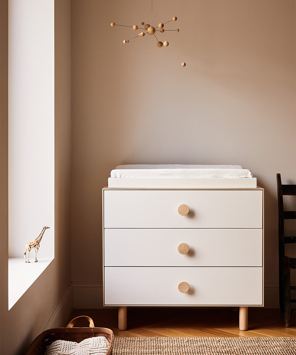 A minimalist nursery features the Dot 3-Drawer Dresser with wooden knobs, a giraffe figurine, and a modern mobile in neutral tones throughout.