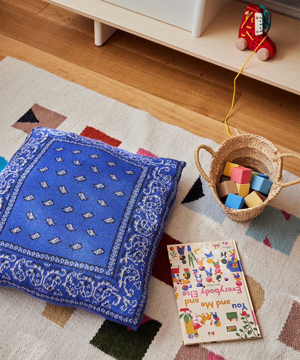 Cozy playroom scene with a Bandana Pillow, woven basket of blocks, colorful book, and toy on a patterned rug.