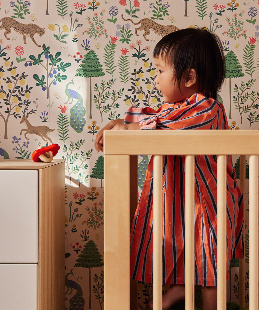 A child in a striped dress holds the Le Baby Bird Rattle while standing by a crib in a room with colorful animal and plant wallpaper.