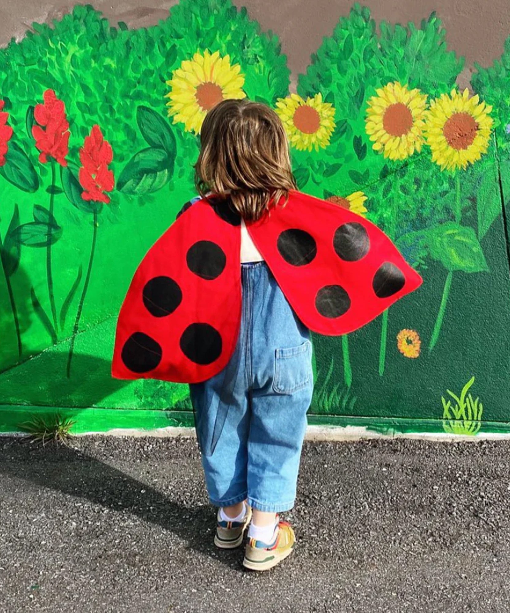 A child wearing Ladybug Wings stands in front of a vibrant mural of flowers and greenery on a wall.