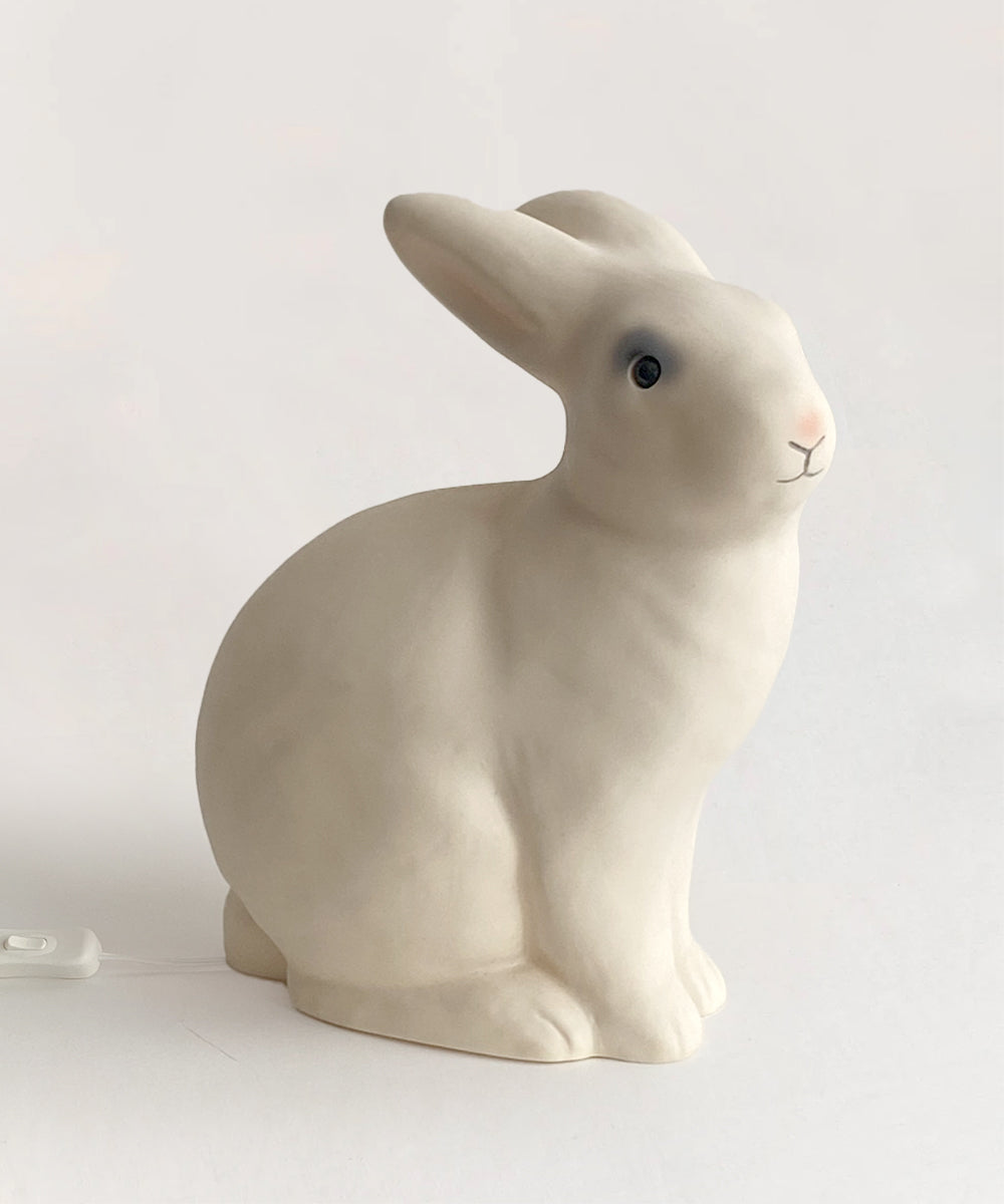 The Le Bunny Lamp, a white rabbit-shaped light featuring a gentle gray accent on one eye, is placed on a light surface and connected to a small white switch.