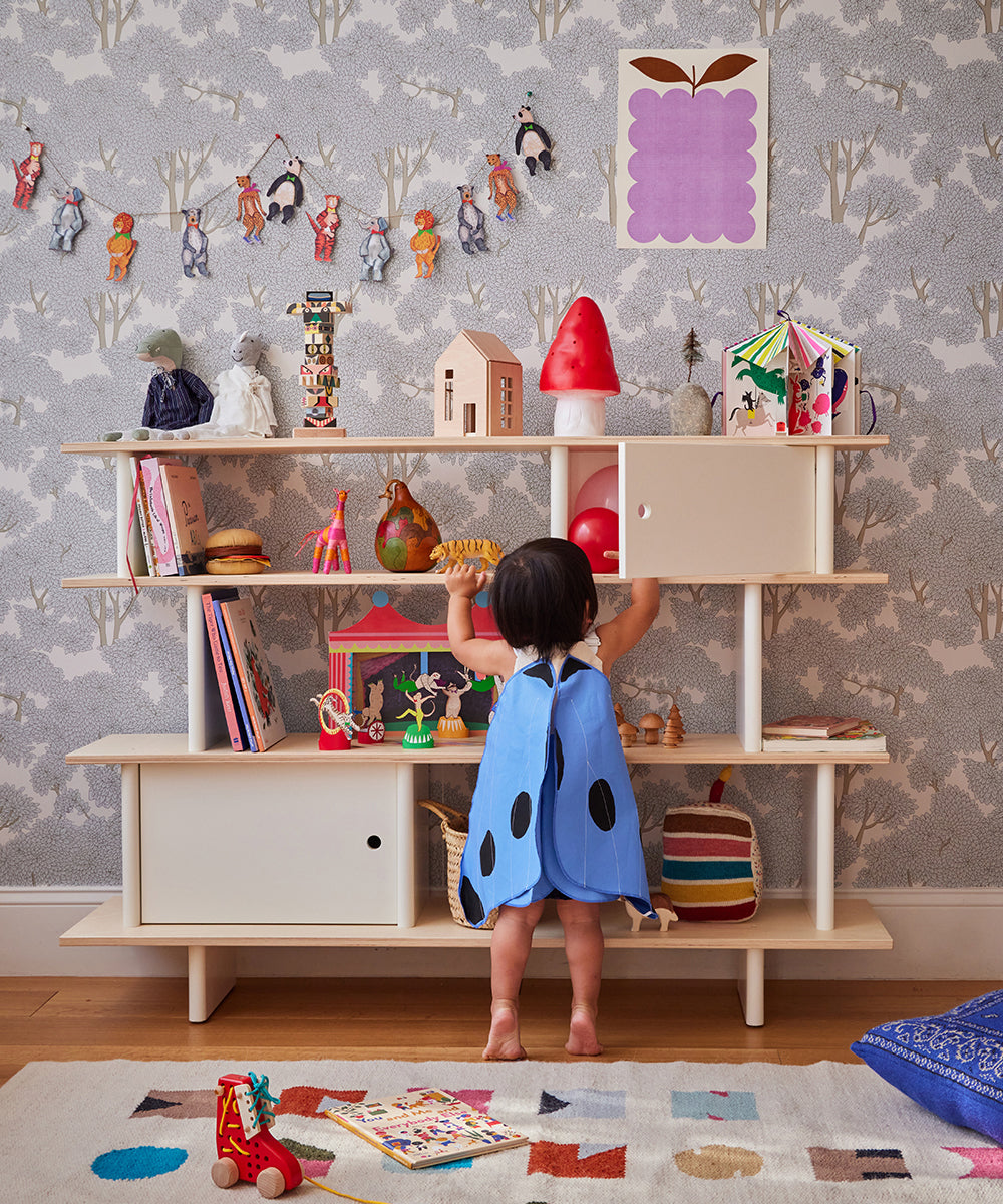 A toddler in a blue dress reaches for toys from the Mini Library on a colorful shelf against patterned wallpaper in the playroom.