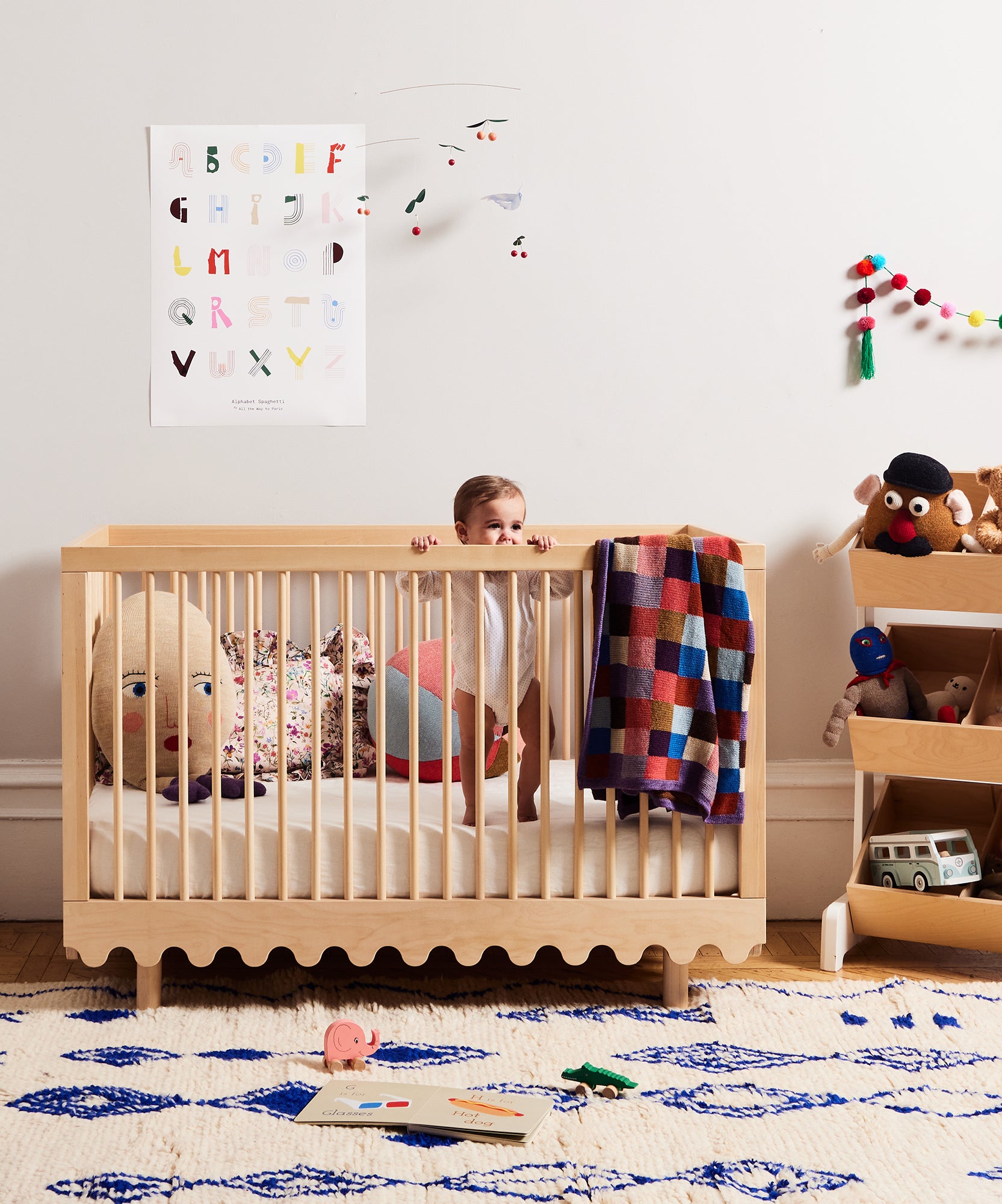 A baby stands in a Moss Crib in a colorful, modern nursery filled with toys and books.