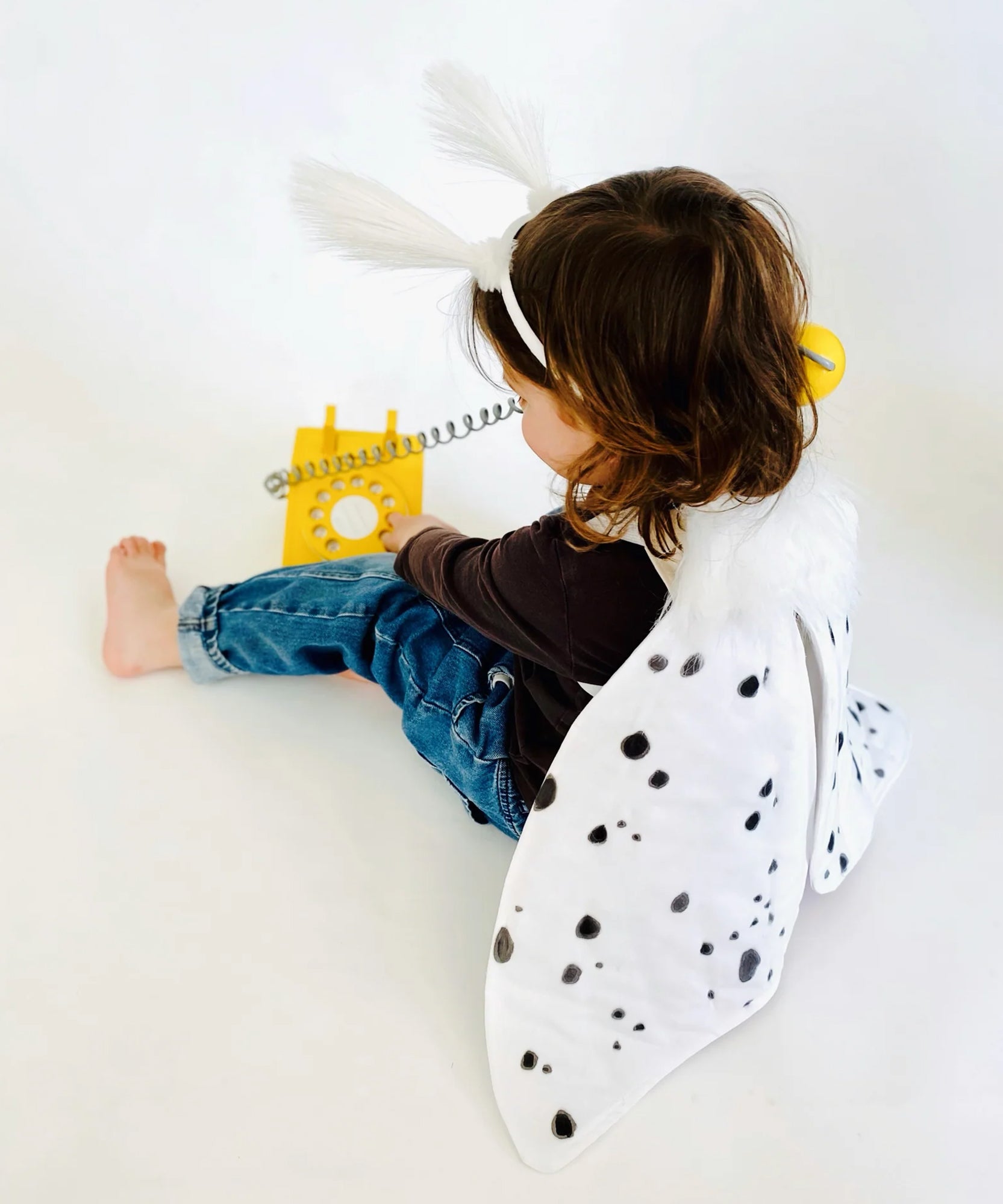 A child wearing Le Leopard Moth Wings cape and headband plays with a yellow rotary phone, sitting on a white floor.