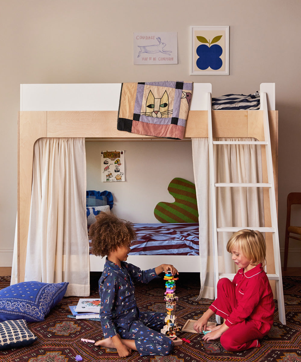 Two children in pajamas play with toys on the floor beside a Bandana Pillow, near a wooden bunk bed with curtains and a ladder.