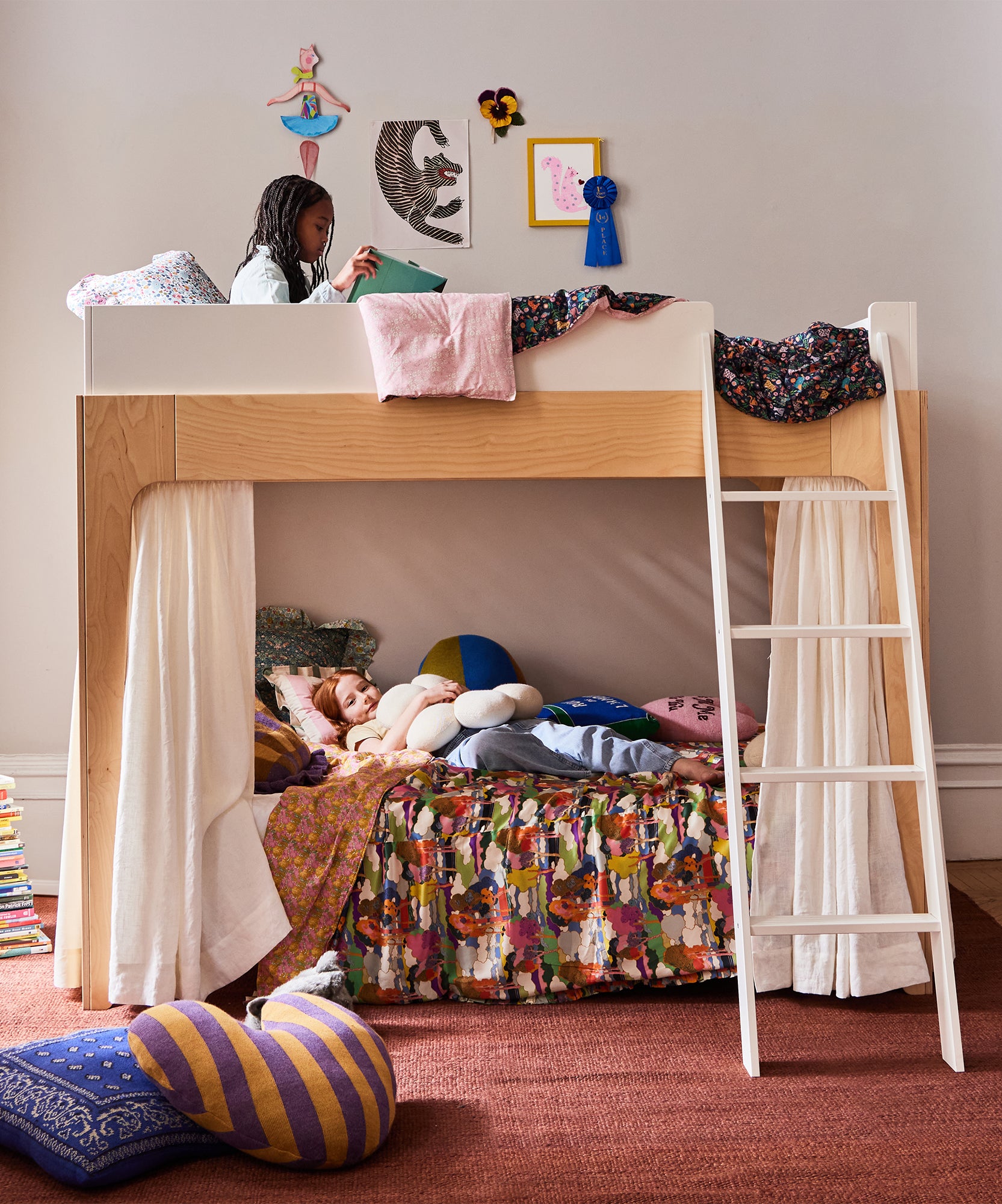 A girl reads on a top bunk with Perch Bunk Bed Curtains, while another child relaxes below in a cozy, decorated kids' room.