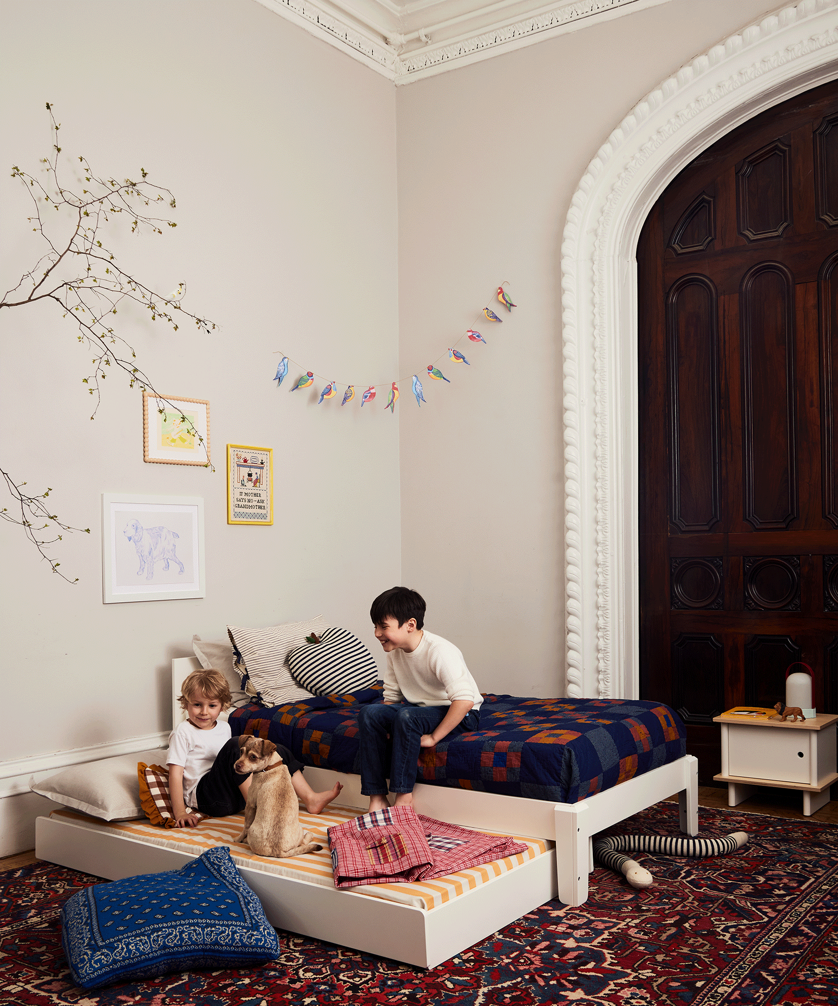 Two children sit on cozy Trundle Mattresses in a stylish bedroom with patterned decor and a large arched wooden door.