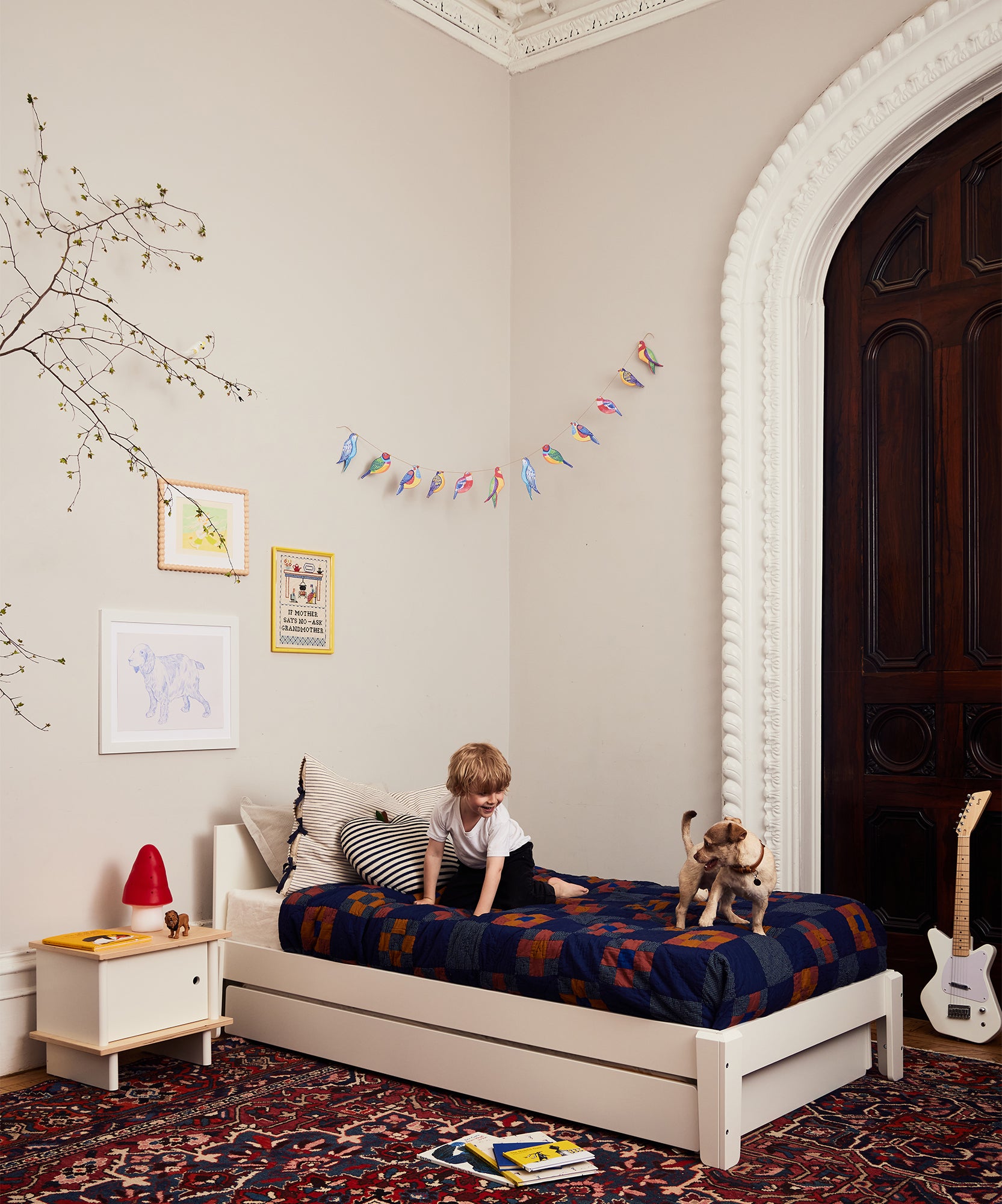 A child sits on a bed with a toy dog in a cozy bedroom decorated with art, books, a guitar, and the Le Small Mushroom Lamp.