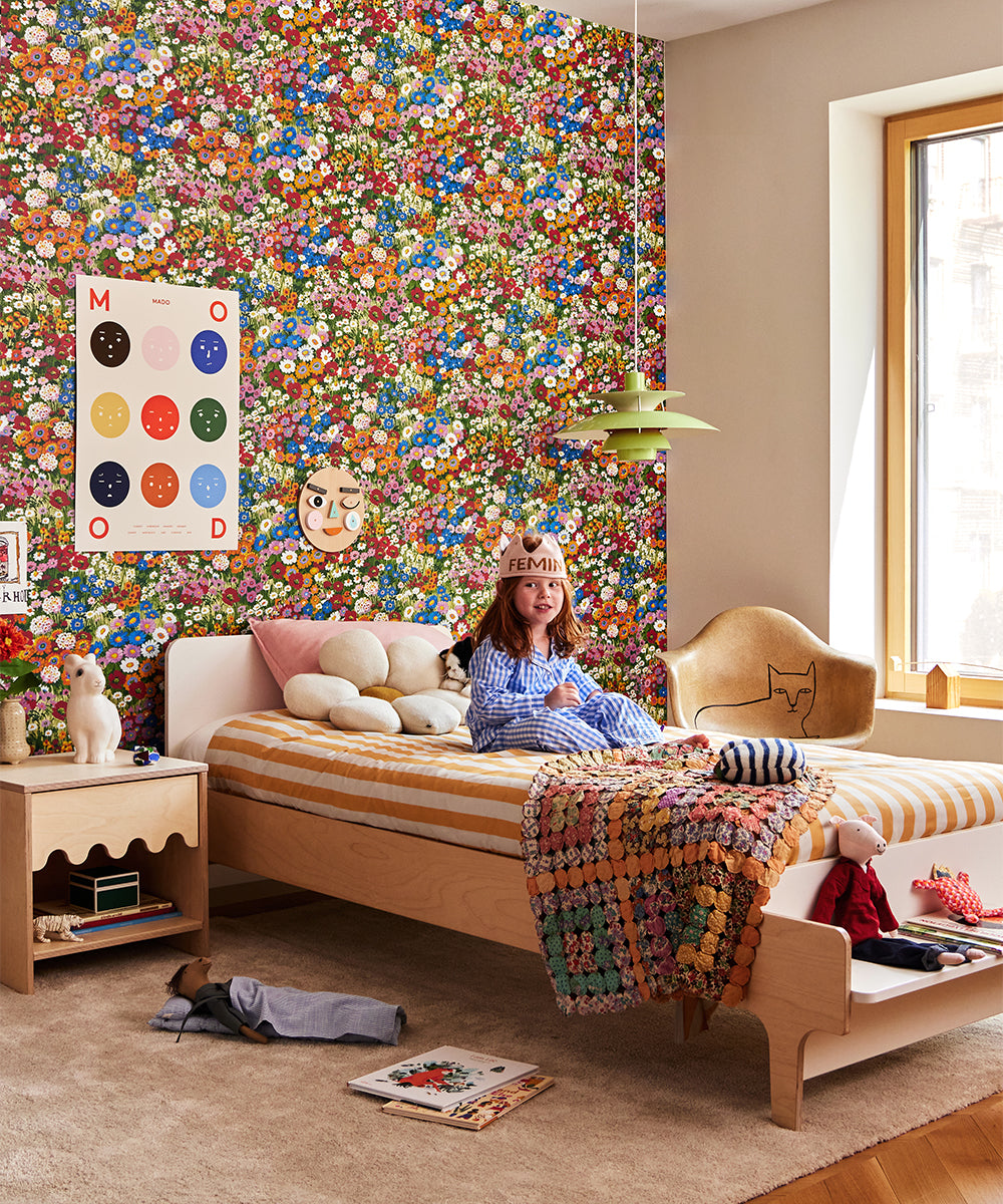 Child with a Daisy Pillow in striped bed, surrounded by colorful floral wallpaper and eclectic decor. Posters on the wall, window on right letting light in.