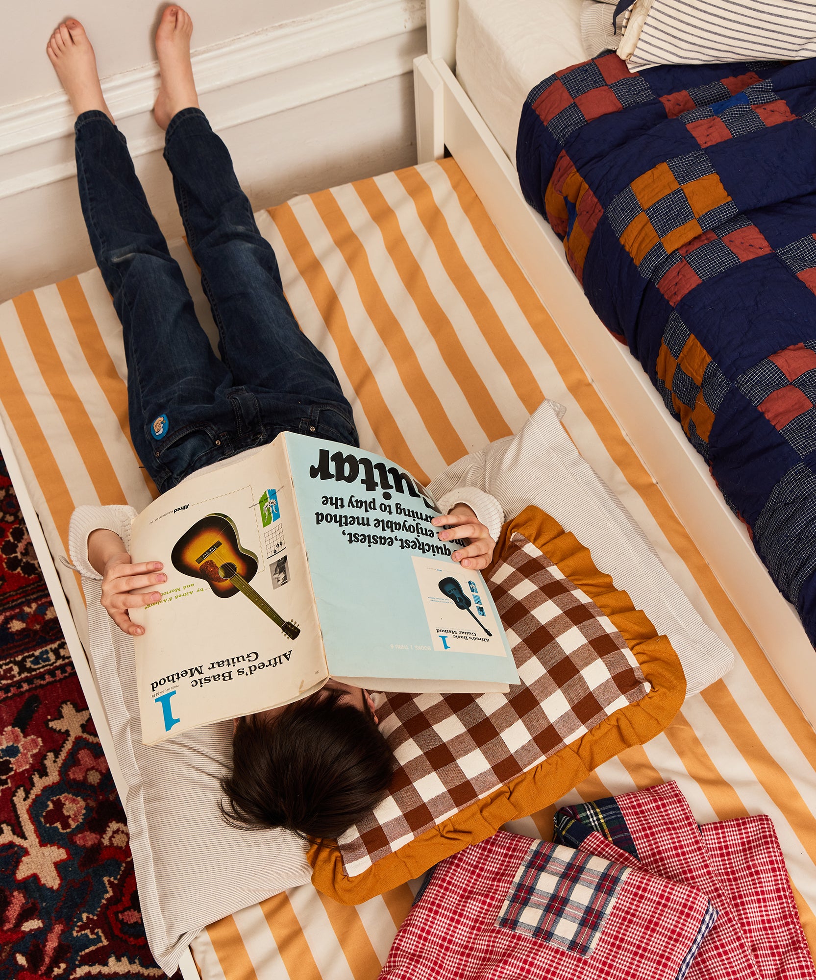A child lies upside down reading on a Trundle Mattress with striped bedding, surrounded by checkered pillows and blankets.
