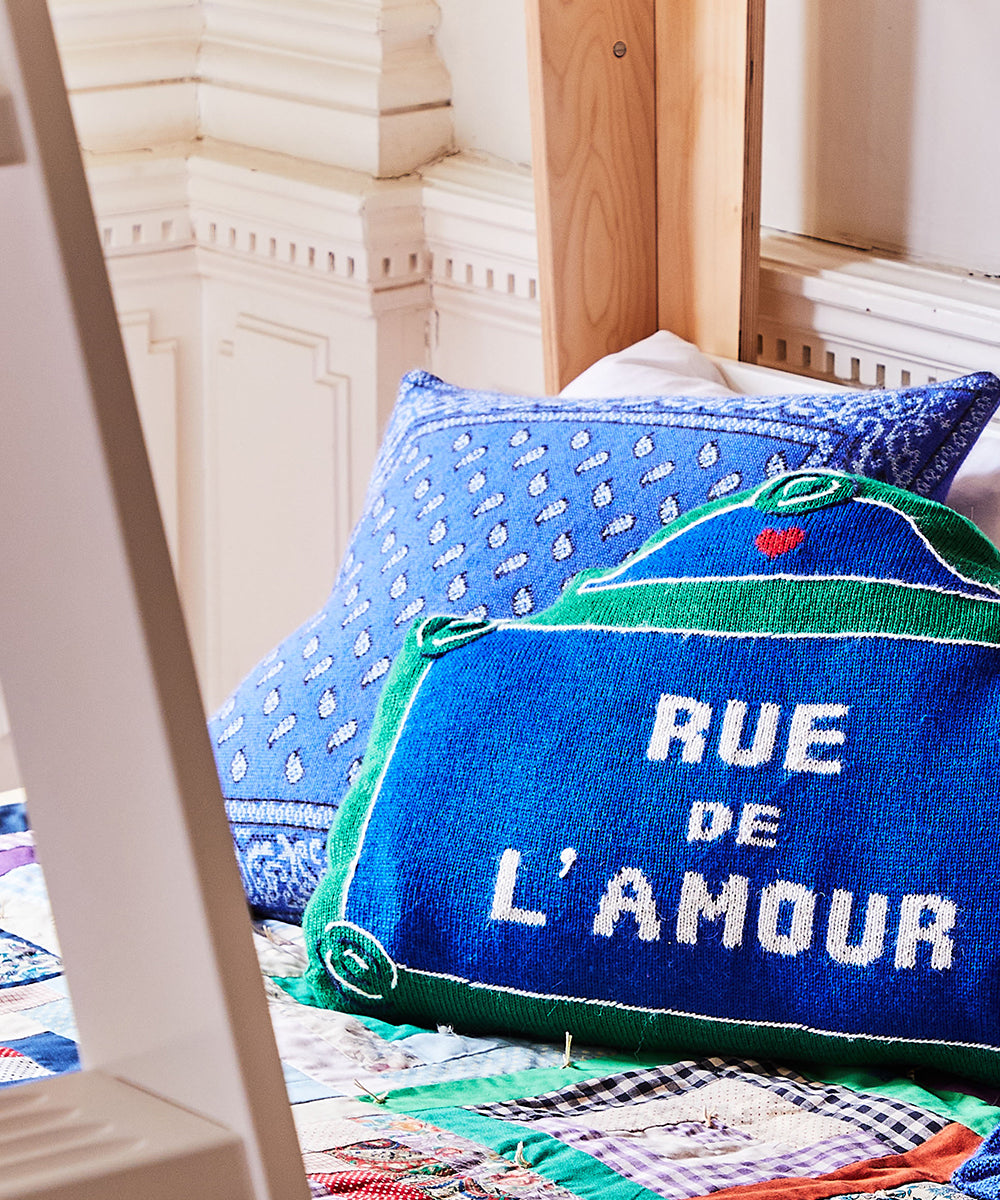 A cozy room features a bed adorned with patterned pillows, including a blue "Rue de L'Amour" Bandana Pillow.
