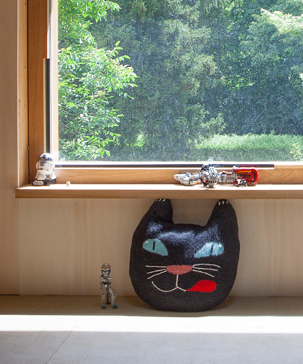 A Black Cat Pillow sits under a sunny window, with toy robots and spaceships lined up on the windowsill.