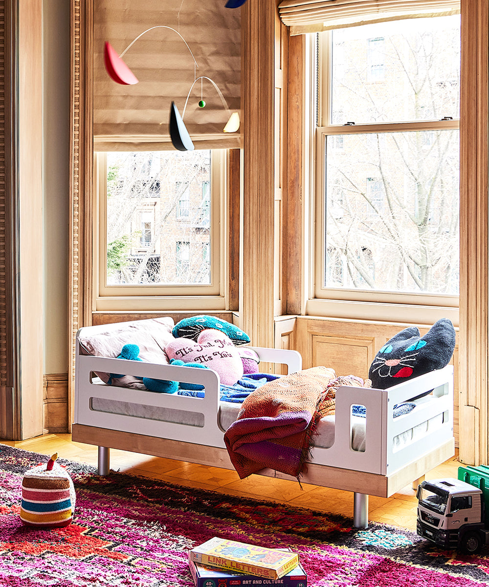 A cozy toddler bed with a Black Cat Pillow sits by large windows in a sunlit room with a vibrant rug.