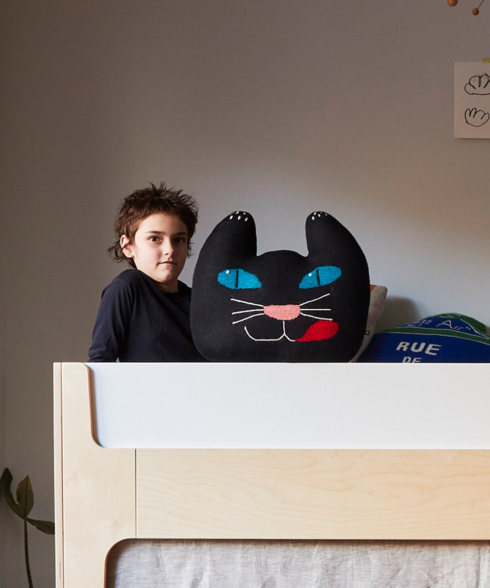 A child sits on a bed beside a Black Cat Pillow featuring bright blue eyes and a red tongue.
