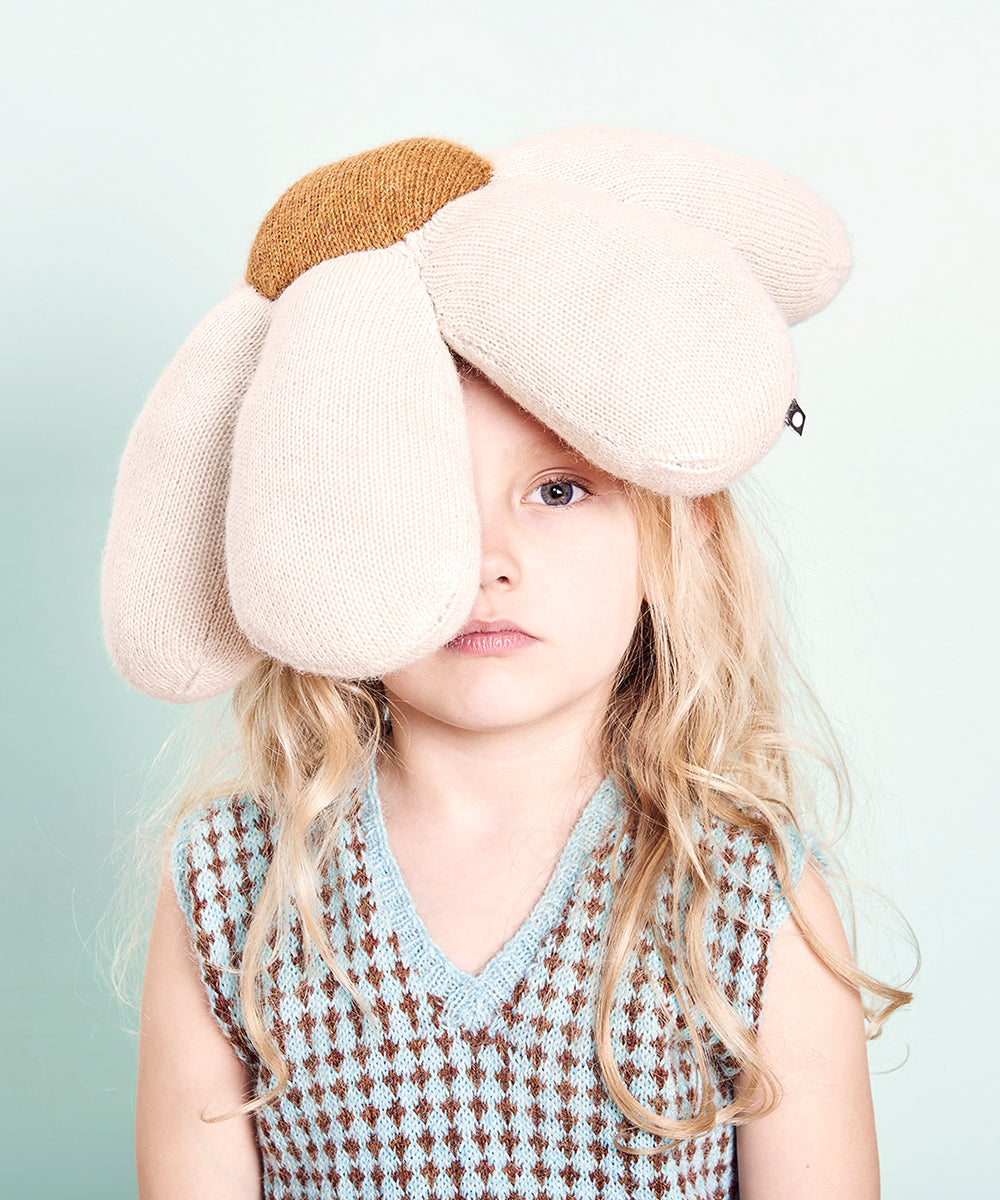 Child in a large Daisy Pillow, knitted like a flower hat over one eye, paired with a patterned vest on a soft green backdrop.